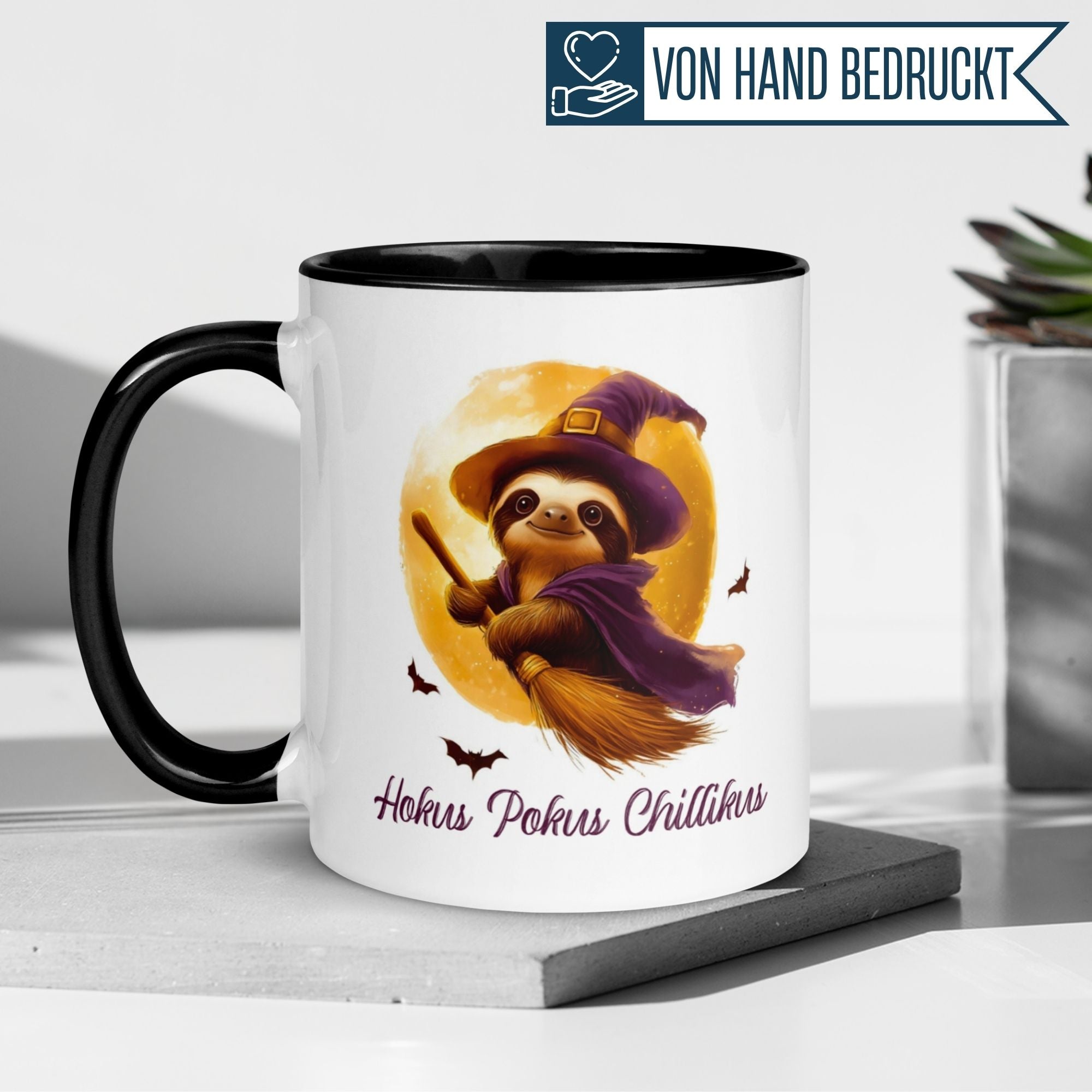 Tasse mit Faultier im Hexenhut und Spruch "Hokus Pokus Chillikus" auf einem Tisch.
