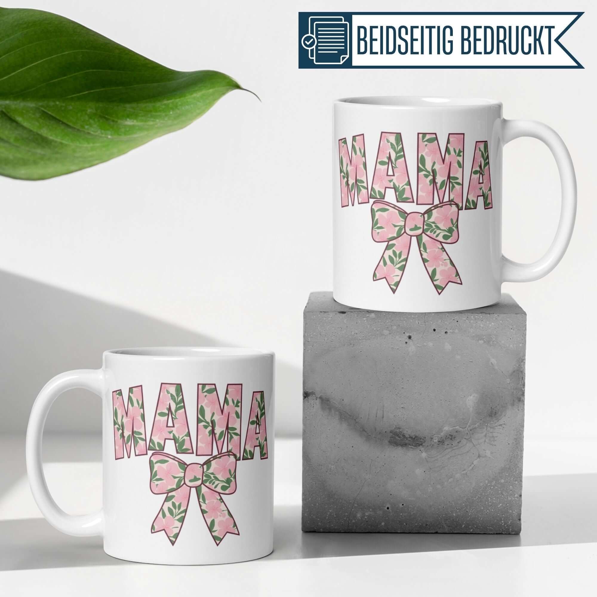 Weißer Becher mit "Mama" Schriftzug und floraler Schleife auf grauem Block, ideal für Geschenke.